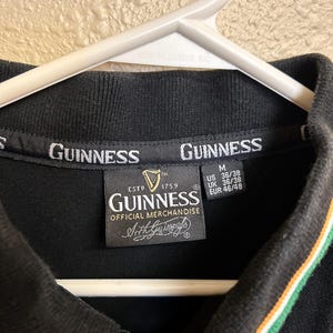 May include: Black polo shirt with the Guinness logo and text. The collar has green and orange stripes. The shirt is labeled as official merchandise and has size information.