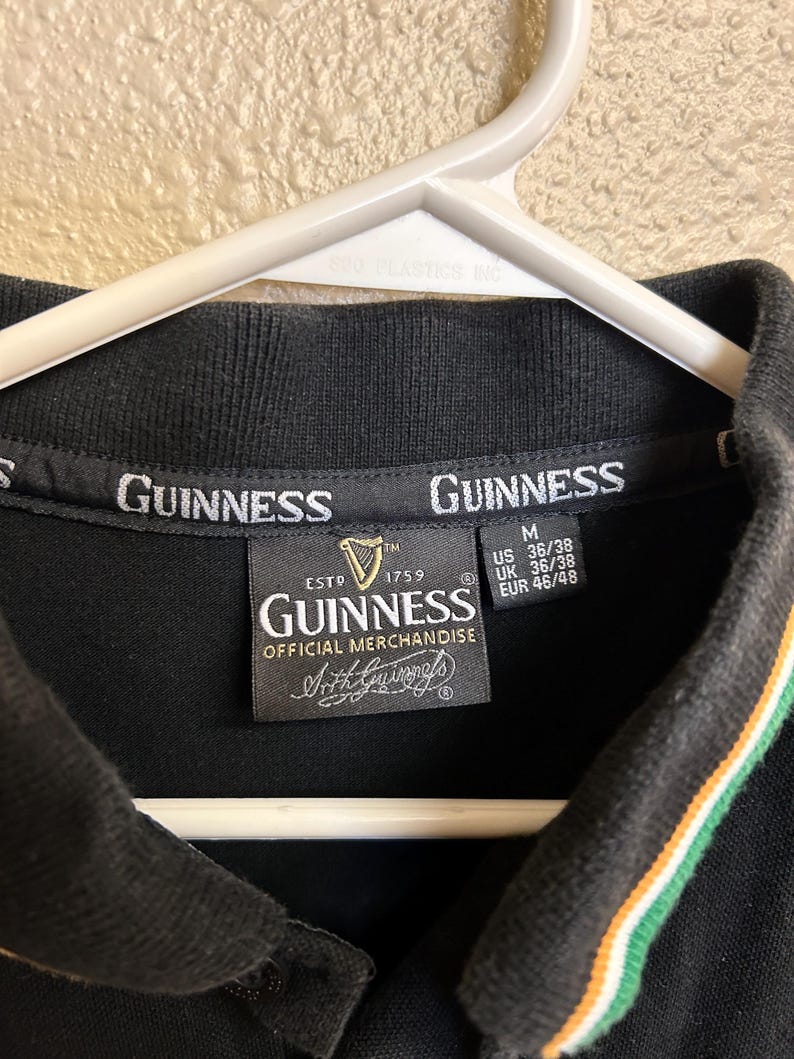 May include: Black polo shirt with the Guinness logo and text. The collar has green and orange stripes. The shirt is labeled as official merchandise and has size information.