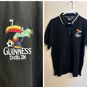 May include: Black polo shirt with a white, green, and red striped collar and sleeve cuffs. The shirt features an embroidered Guinness Dublin logo with a toucan, soccer ball, and the word "Ireland".
