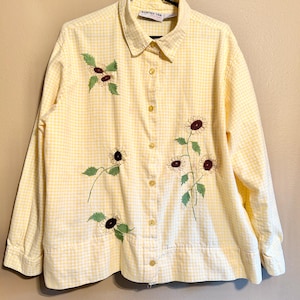 May include: Yellow gingham button-down shirt with long sleeves. The shirt features embroidered sunflowers with green leaves and maroon centers. The shirt is hanging on a black hanger.
