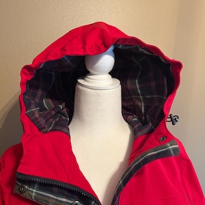 Puede incluir: Chaqueta con capucha de color rojo brillante con forro de cuadros visible en la capucha y el cuello. La chaqueta tiene cierre de cremallera y detalles de botones a presi&oacute;n. La capucha est&aacute; levantada y la chaqueta se muestra en un maniqu&iacute; blanco.