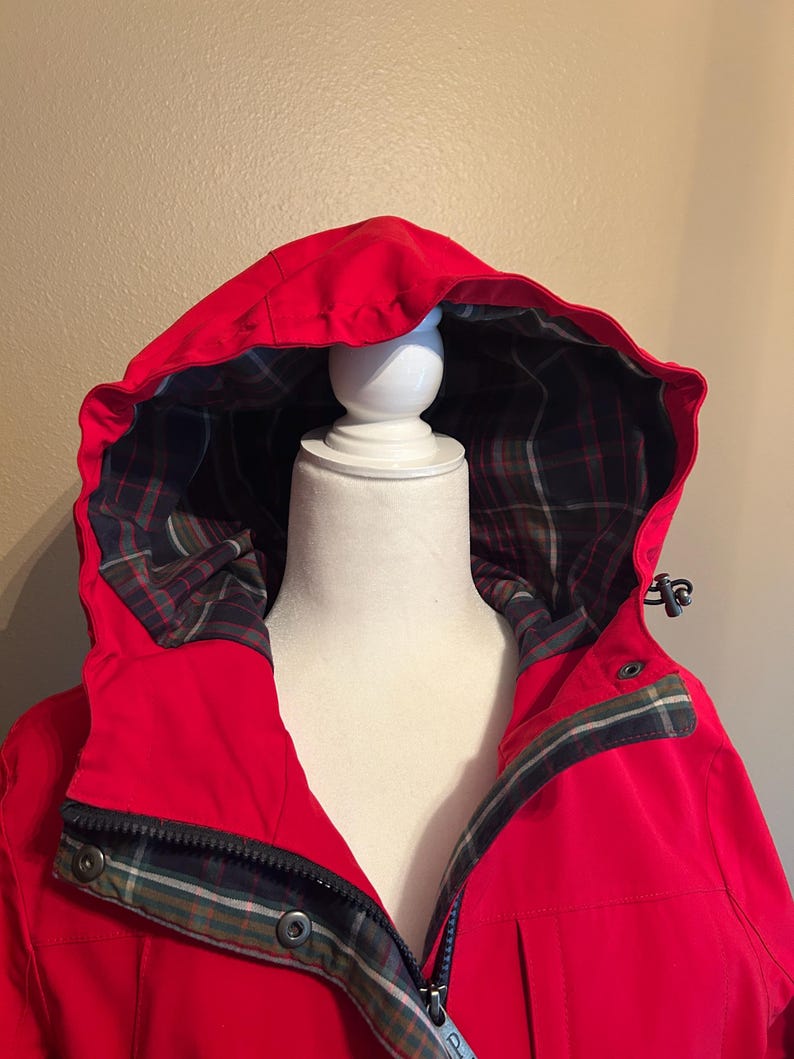 Puede incluir: Chaqueta con capucha de color rojo brillante con forro de cuadros visible en la capucha y el cuello. La chaqueta tiene cierre de cremallera y detalles de botones a presi&oacute;n. La capucha est&aacute; levantada y la chaqueta se muestra en un maniqu&iacute; blanco.