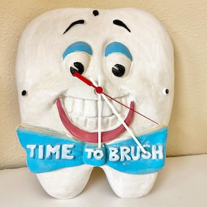 May include: A white wall clock shaped like a smiling tooth with blue eyebrows and a blue bowtie. The bowtie features the text TIME TO BRUSH in white letters. The clock has black eyes, a red mouth, and three clock hands in red and white colors.