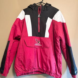 May include: A red, black, and white Arizona Cardinals anorak jacket. The jacket has a hood, a front pocket, and a zipper. The team logo is on the front pocket. The jacket is hanging on a black hanger.