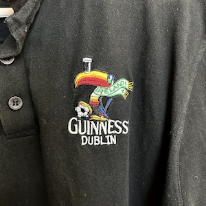 May include: Black polo shirt with a Guinness Dublin logo featuring a toucan with a pint of Guinness on its head, a soccer ball, and an Ireland scarf. The collar has green, white, and orange stripes.