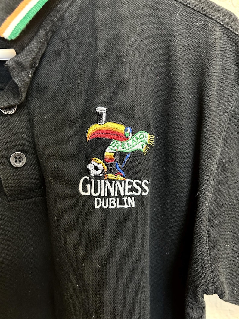 May include: Black polo shirt with a Guinness Dublin logo featuring a toucan with a pint of Guinness on its head, a soccer ball, and an Ireland scarf. The collar has green, white, and orange stripes.