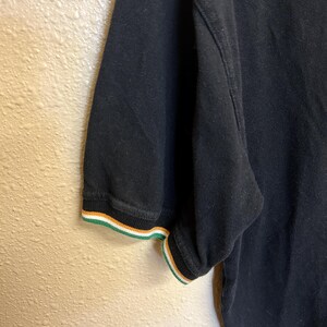 May include: Black polo shirt with short sleeves. The sleeve cuff features a striped design in green, white, and orange. The shirt is made of a textured fabric and is partially visible.