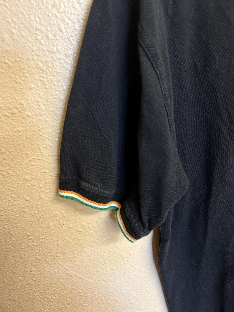 May include: Black polo shirt with short sleeves. The sleeve cuff features a striped design in green, white, and orange. The shirt is made of a textured fabric and is partially visible.