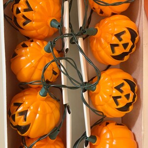 May include: A string of orange plastic jack-o'-lantern lights with black cut-out faces. The lights are strung on a green wire and are arranged in a box. A festive Halloween decoration.