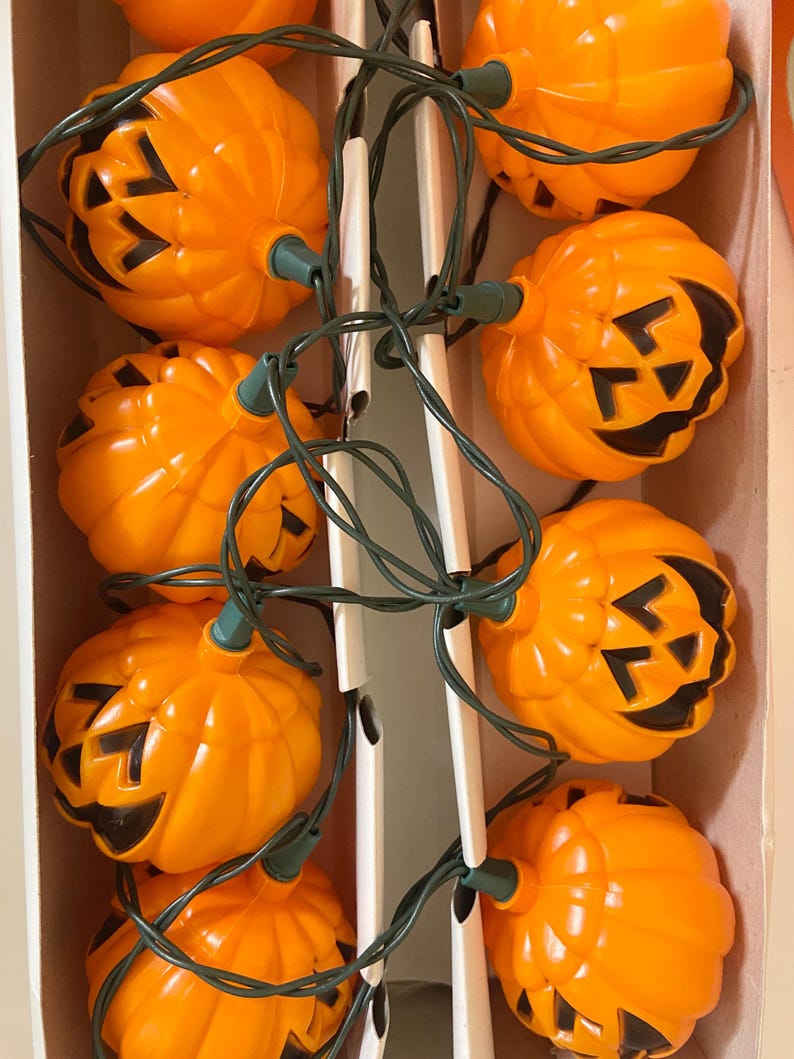 May include: A string of orange plastic jack-o'-lantern lights with black cut-out faces. The lights are strung on a green wire and are arranged in a box. A festive Halloween decoration.