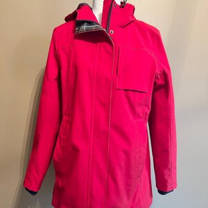 Puede incluir: Chaqueta roja brillante con capucha y cierre de cremallera. La chaqueta tiene un bolsillo en la parte delantera y un forro a cuadros en la capucha. Las mangas tienen pu&ntilde;os azul oscuro. La chaqueta se exhibe en un maniqu&iacute; blanco.