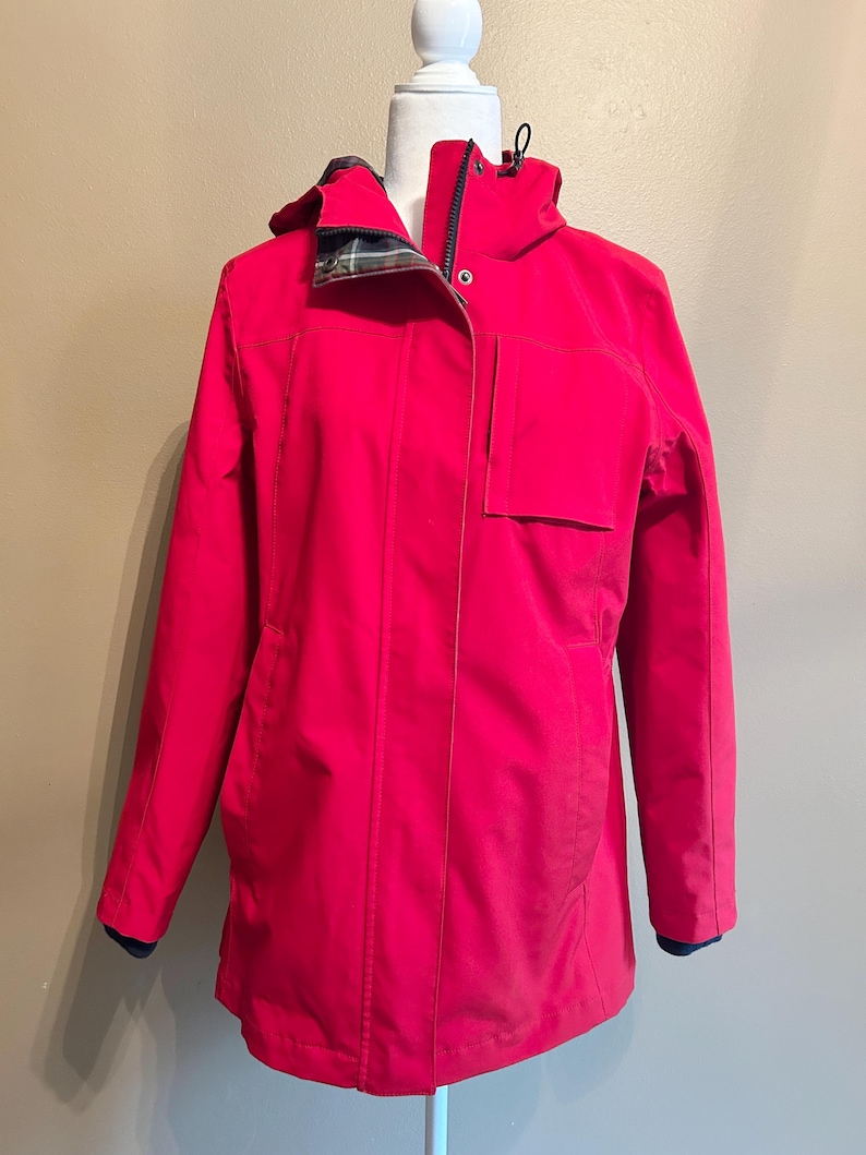 Puede incluir: Chaqueta roja brillante con capucha y cierre de cremallera. La chaqueta tiene un bolsillo en la parte delantera y un forro a cuadros en la capucha. Las mangas tienen pu&ntilde;os azul oscuro. La chaqueta se exhibe en un maniqu&iacute; blanco.