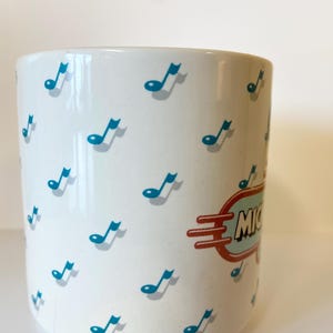 Pode incluir: Caneca de cer&acirc;mica branca com um padr&atilde;o de notas musicais azuis e uma placa retr&ocirc; vermelha e azul que diz "Michigan".