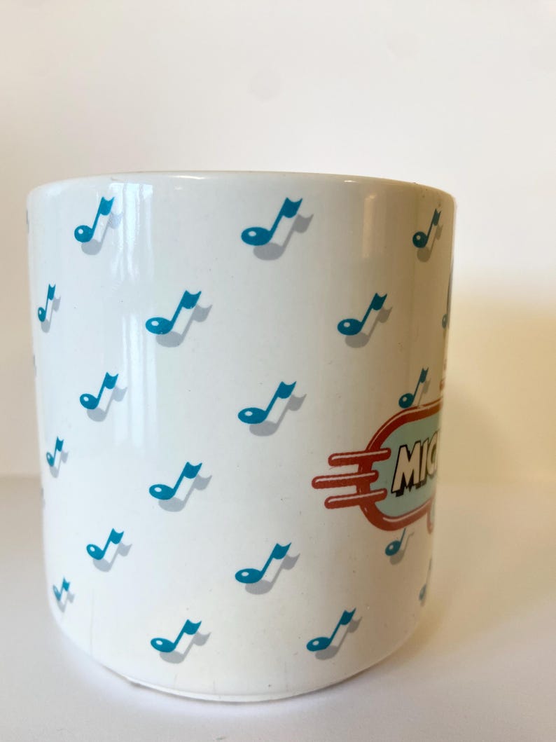 Pode incluir: Caneca de cer&acirc;mica branca com um padr&atilde;o de notas musicais azuis e uma placa retr&ocirc; vermelha e azul que diz "Michigan".