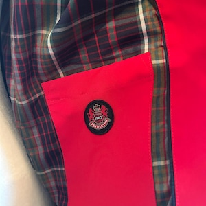 Puede incluir: Primer plano de una chaqueta roja con forro a cuadros en tonos verdes, marrones y negros. Se ve un bolsillo rojo, con un emblema circular que dice "Pendleton 1863". La chaqueta tiene una cremallera negra.