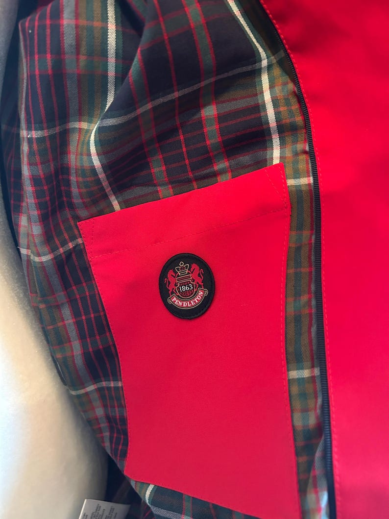 Puede incluir: Primer plano de una chaqueta roja con forro a cuadros en tonos verdes, marrones y negros. Se ve un bolsillo rojo, con un emblema circular que dice "Pendleton 1863". La chaqueta tiene una cremallera negra.