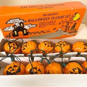 May include: An orange box labeled "Decorative Halloween 10 Light Set" with a string of orange pumpkin lights. The box features Halloween-themed illustrations of a haunted house, ghosts, and a jack-o'-lantern. The lights are in a white cardboard tray.