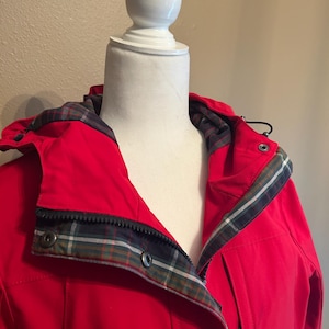 Puede incluir: Una chaqueta roja brillante con capucha y forro de cuadros visible alrededor del cuello y la capucha. La chaqueta tiene cierre de cremallera y botones de presi&oacute;n. La chaqueta se exhibe en un maniqu&iacute; blanco.