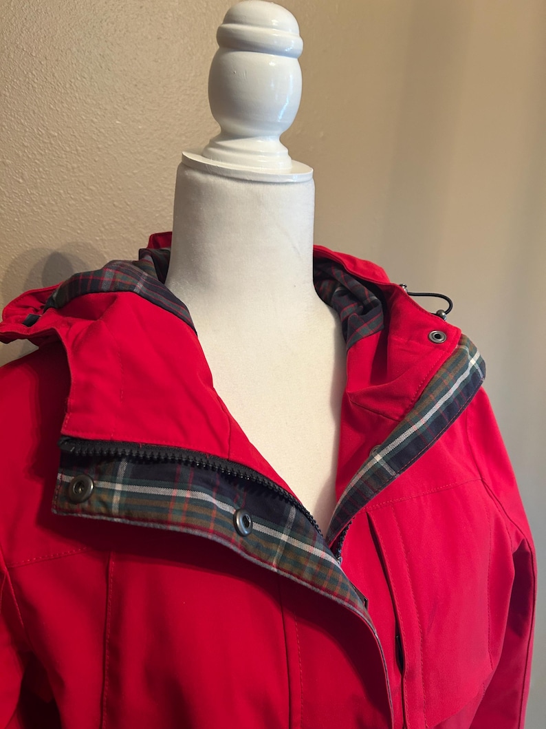 Puede incluir: Una chaqueta roja brillante con capucha y forro de cuadros visible alrededor del cuello y la capucha. La chaqueta tiene cierre de cremallera y botones de presi&oacute;n. La chaqueta se exhibe en un maniqu&iacute; blanco.