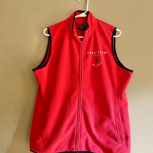 May include: A red fleece vest with a full zipper and a stand-up collar. The vest has black trim along the edges and a small logo on the chest. The vest is hanging on a white hanger.