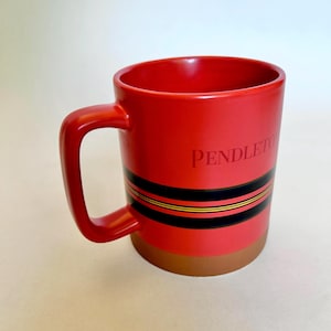 May include: A red ceramic mug with a handle, featuring the text PENDLETON printed in red. The mug has a horizontal black and gold striped pattern around the center and a solid brown base. The interior and exterior are finished in a smooth red glaze.
