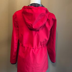 Puede incluir: Una chaqueta roja brillante con capucha y mangas largas. La chaqueta tiene una cintura con cord&oacute;n y un forro a cuadros visible en la abertura de la capucha. Los pu&ntilde;os tienen un ribete azul marino. La chaqueta se muestra en una cabeza de maniqu&iacute; blanca.