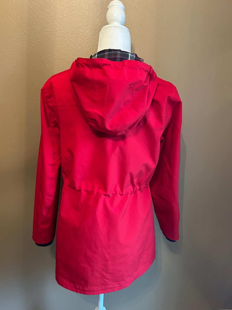 Puede incluir: Una chaqueta roja brillante con capucha y mangas largas. La chaqueta tiene una cintura con cord&oacute;n y un forro a cuadros visible en la abertura de la capucha. Los pu&ntilde;os tienen un ribete azul marino. La chaqueta se muestra en una cabeza de maniqu&iacute; blanca.