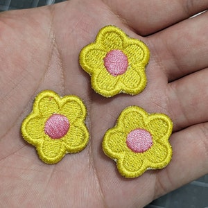 Puede incluir: Tres parches de flores de fieltro amarillo con centros rosas. Las flores tienen un diámetro de aproximadamente 2,5 cm.