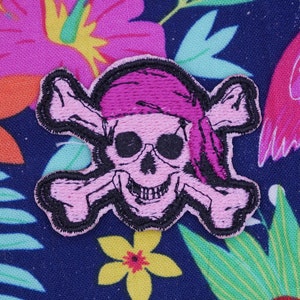 May include: Pink and black fabric pirate skull and crossbones patch with a pink bandana on the skull. The patch is on a blue background with a pink flamingo and tropical flowers.
