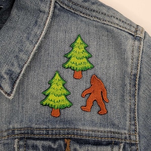Może przedstawiać: Dżinsowa kurtka z trzema naszywkami: dwoma zielonymi drzewami i brązowym Yeti.