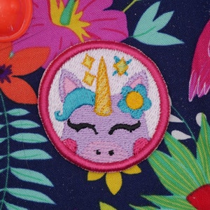 May include: An embroidered patch featuring a unicorn design in pink and purple. The unicorn has a gold horn, a yellow flower, and its eyes are closed. The patch is edged with a pink border.