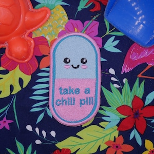 Op de afbeelding: Een blauwe en roze pilvormige patch met een glimlachend gezicht en de tekst "take a chill pill". De patch bevindt zich op een donkerblauwe stof met een tropische print met ananas, bloemen en palmbladeren.