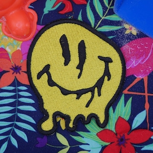 Könnte beinhalten: Ein gelber Smiley-Patch mit einem Tropfeffekt auf blauem Hintergrund mit tropischem Blumendruck. Der Patch hat einen schwarzen Umriss und ein schwarzes Lächeln.