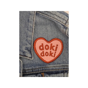 Puede incluir: Un parche en forma de corazón rosa con el texto "doki doki" bordado en rojo.
