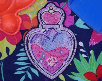 May include: A pink and purple heart-shaped bottle with a pink lid and glitter accents. The bottle is embroidered with a floral design and is surrounded by a tropical print.
