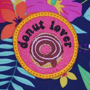 May include: A pink and yellow embroidered patch with the text "Donut Lover" and a brown and white donut design.