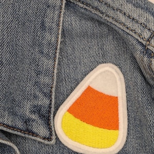 Pode incluir: Um remendo de feltro branco em forma de milho doce com bordados laranja e amarelo. O remendo é costurado em uma jaqueta jeans azul.