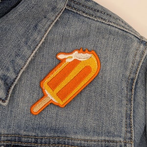 Może przedstawiać: Naszywka z wyszywaną pomarańczową i żółtą lodową pałeczką na niebieskiej dżinsowej kurtce.