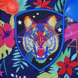 Puede incluir: Un parche bordado colorido con un tigre en un esquema de color azul, amarillo y rojo. El tigre está sobre un fondo negro con estrellas blancas. El parche está rodeado por un borde azul.