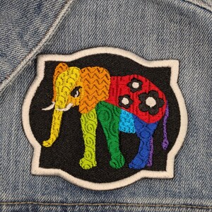 May include: A rainbow-coloured embroidered elephant patch with black outlines and white stitching. The patch is on a blue denim jacket.