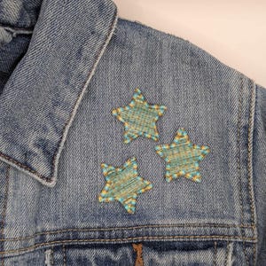 May include: A blue denim jacket with three embroidered stars on the front. The stars are made of yellow and blue thread and have a checkered pattern.