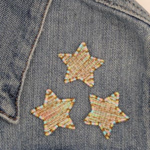 May include: Three embroidered stars in shades of brown, pink, and blue are sewn onto a denim jacket.