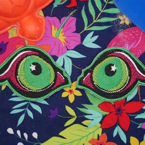 May include: Two green and black embroidered eye patches with a white star in the center of each eye. The patches are on a blue background with pink and green floral designs.