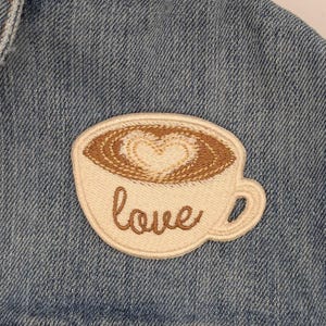 Op de afbeelding: Een beige geborduurde patch met een bruin koffiekopje ontwerp en het woord "love" in bruin borduurwerk. De patch is genaaid op een blauw denim jack.