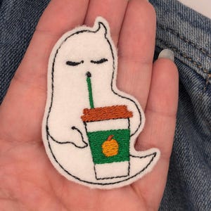 May include: White ghost with closed eyes holding a green and white coffee cup with an orange pumpkin on it. The cup has a brown lid and a straw.