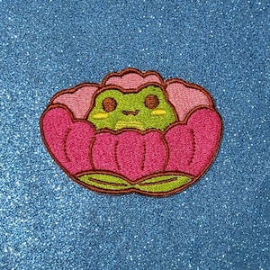 Puede incluir: Una rana verde con ojos marrones y una sonrisa está sentada dentro de una flor rosa con hojas verdes. La flor está bordada sobre un fondo azul brillante.