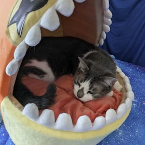 May include: A gray and white cat curled up inside a novelty pet bed shaped like a cartoon mouth. The bed is yellow with white teeth-shaped trim and a red interior. The cat is sleeping on a red cushion.