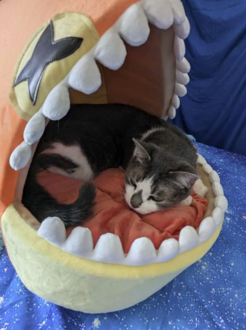 May include: A gray and white cat curled up inside a novelty pet bed shaped like a cartoon mouth. The bed is yellow with white teeth-shaped trim and a red interior. The cat is sleeping on a red cushion.