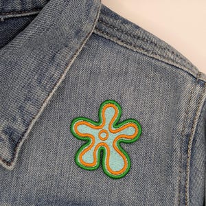 May include: A denim jacket with a colorful embroidered flower patch. The patch features a light blue center, outlined in orange, with a green border. The jacket is a light blue wash.
