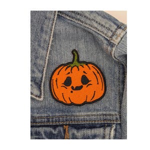 May include: An orange pumpkin patch with a green stem and black facial features is sewn onto a denim jacket. The patch has a black outline and a smiling expression, perfect for Halloween or fall-themed apparel.